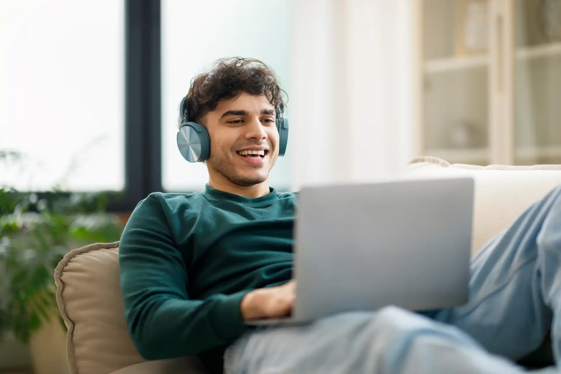 A young man with dark curly hair wearing headphones and a green shirt smiles while lounging on a couch with his laptop computer.