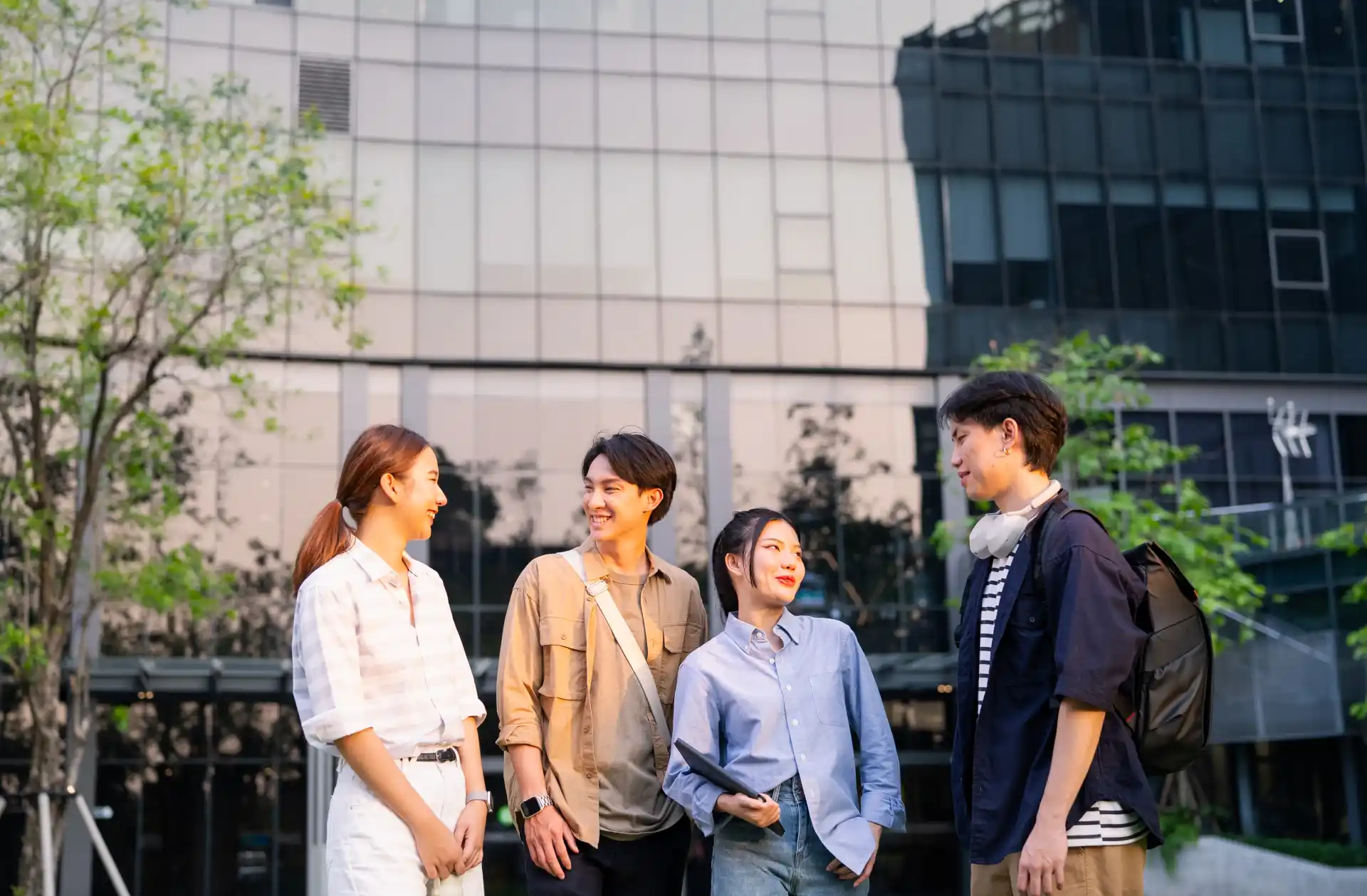 A group of Asian college students in button up shirts stand outside talking and smiling in front of a large glass building.