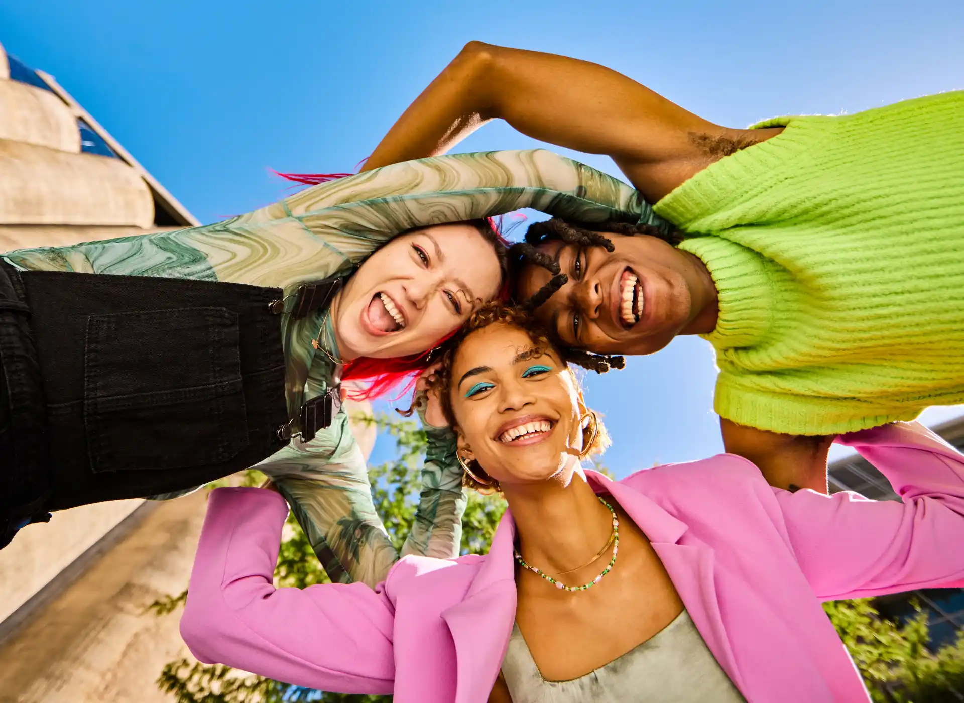 Two black young adults and a young Asian woman with red hair lock arms in a huddle smiling and laughing.