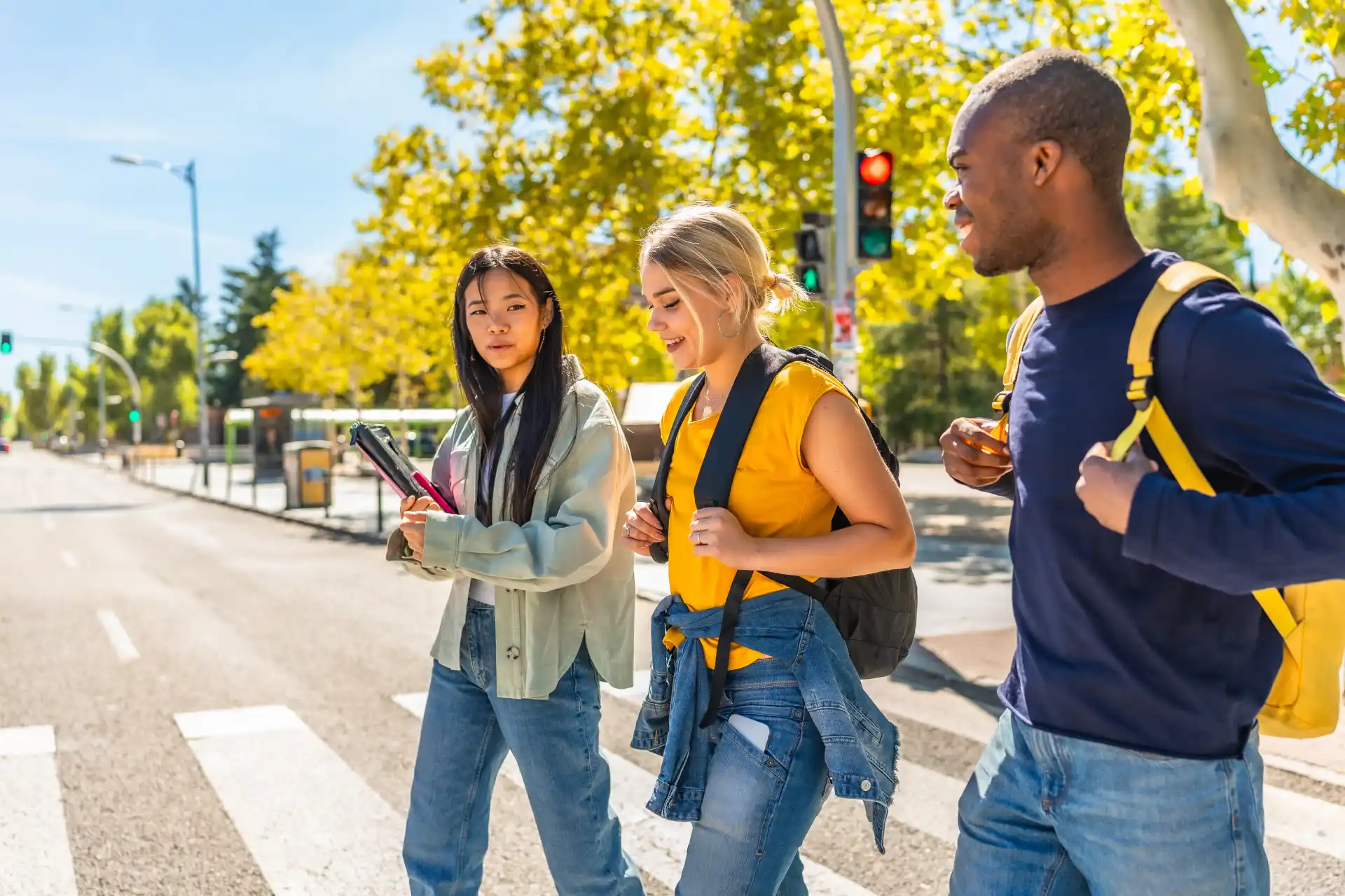 Two young women and a young man wearing backpacks chat while crossing a crosswalk on a sunny day.