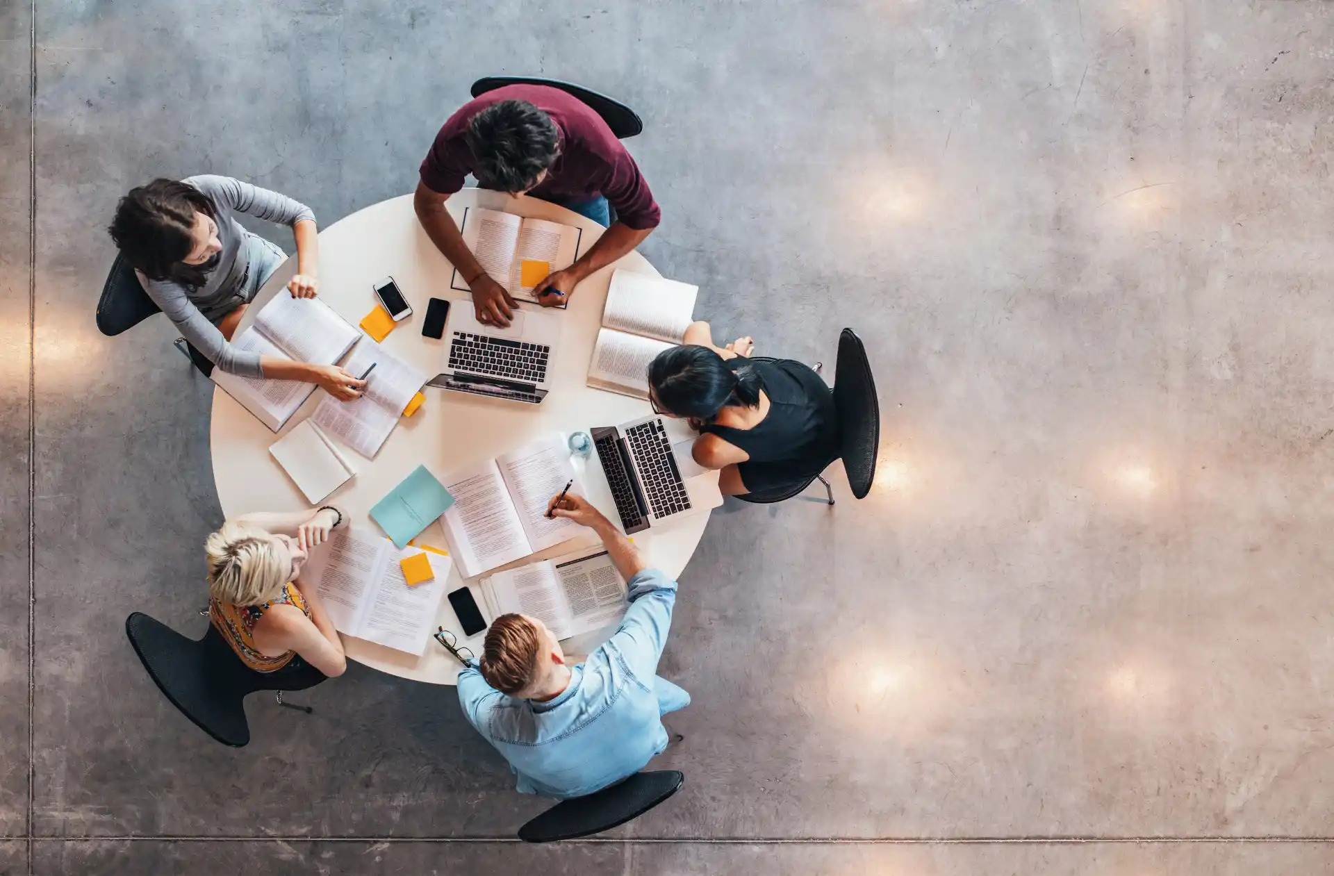 birds eye view of five students sitting at a round white study table with various books and laptops open.